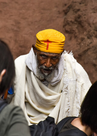 A man in traditional religious attire, including a yellow hat with a cross, stands indoors in Lalibela, Ethiopia.