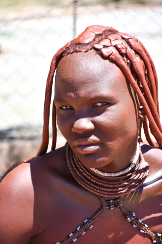 A Himba woman with traditional hair and necklaces gazes at the camera in a close up portrait.