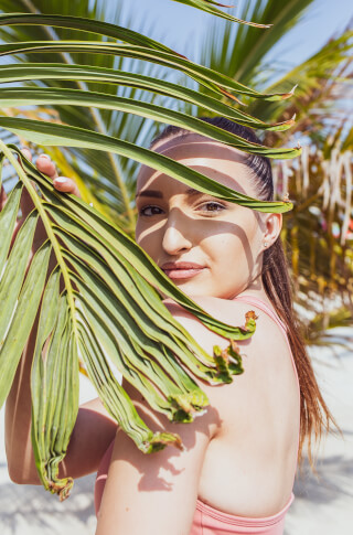 A woman with brown hair poses with a palm leaf on a sunny beach.