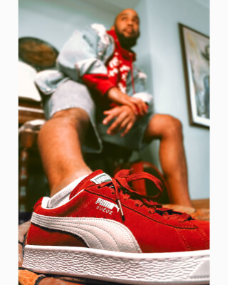 A man poses with red Puma Suede shoes in a studio setting, showcasing vibrant streetwear style.