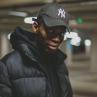 A stylish man in a black puffer jacket and NY Yankees cap poses in an urban parking garage setting.