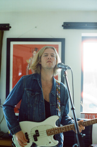 A musician plays a white bass guitar and sings into a microphone in a home studio.