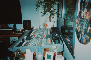 A mixing console is on a desk with a clock, plant, and other equipment in a music studio.