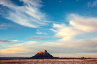A mesa rises from a flat, expansive landscape under a blue sky with streaking clouds in a scenic outdoor view.