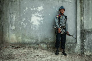 A soldier in uniform stands guard against a concrete wall, armed with a rifle.