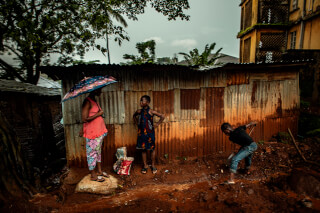 Three people are outside a corrugated metal structure in a developing urban environment.