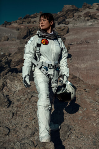 An astronaut walks on a rocky, desolate, Mars-like planet wearing a white spacesuit and carrying a helmet.