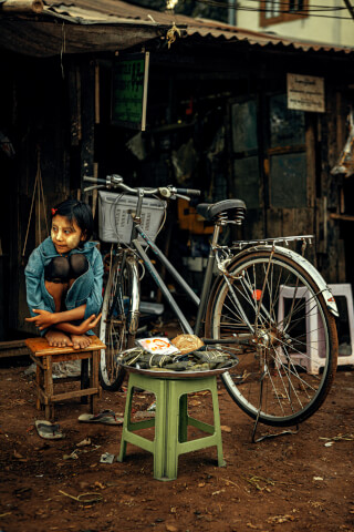 A young girl with thanaka sits beside a bicycle and food stand in an outdoor setting.