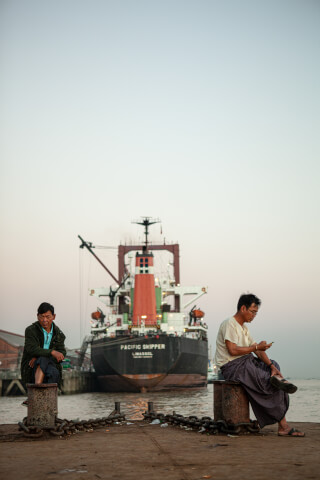 Two men sit on a pier near a large cargo ship at sunset, capturing a moment of quiet contemplation.