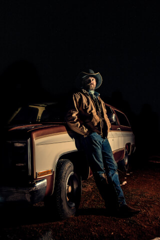 A man in a cowboy hat leans against a vintage truck at night in a rural setting.