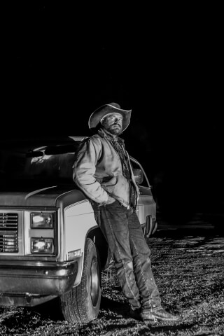 A cowboy stands next to his truck at night in a black and white portrait.