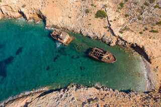 Aerial view of two old shipwrecks in a secluded bay with clear turquoise water, surrounded by rocky coastline.