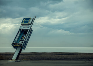 An abandoned and vandalized phone booth tilts precariously on a beach against a cloudy sky.