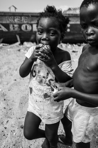 A black and white portrait of a child drinking from a bottle on the beach near another child.