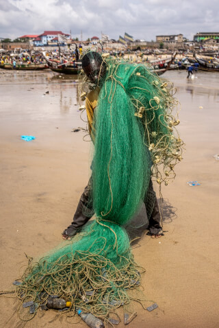 A fisherman carries a large green fishing net on the beach in a coastal community.