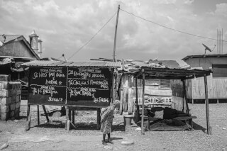 A grayscale shot captures a chalkboard with soccer scores in a modest street setting.