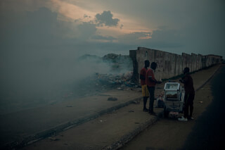 Three men stand by a food cart next to a landfill and smoke in a developing country, highlighting poverty and environmental issues.