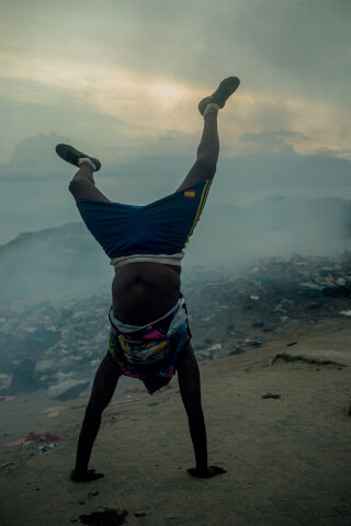A boy performs a handstand at a dump, highlighting themes of poverty and resilience.