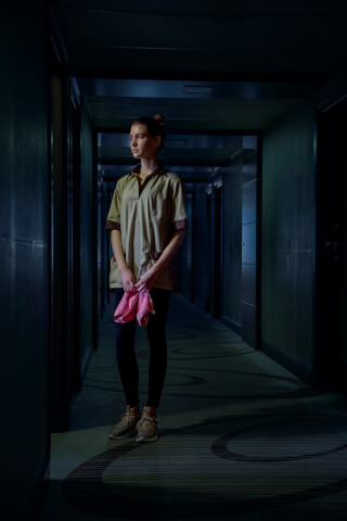 A maid stands in a dimly lit hotel hallway, holding cleaning cloths.