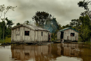 Wooden houses stand on stilts above the river in a rural landscape with people and a boat.