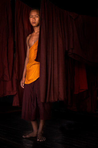 A young monk stands amidst hanging robes, creating a scene of quiet contemplation and cultural immersion.