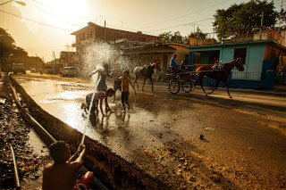 A group of children play with water on a street in Cuba as a horse-drawn vehicle passes by during a bright, sunny day.