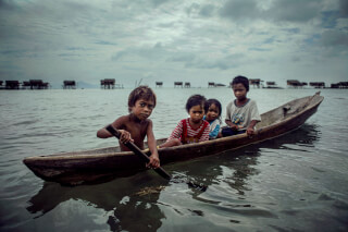 Four children are in a small boat on the water, with houses on stilts in the background under a cloudy sky.