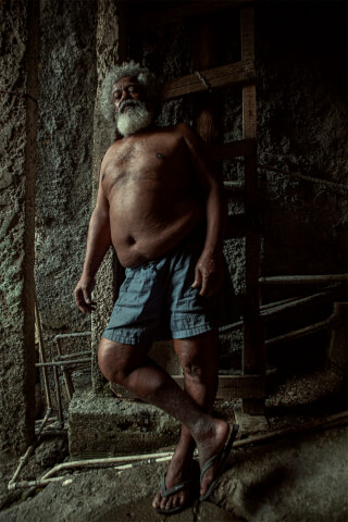An older man with a beard stands indoors in shorts and flip flops.