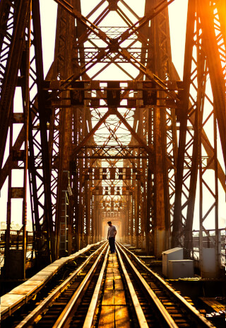 A man walks on a railway bridge, framed by its intricate steel structure in warm sunlight.