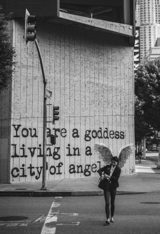 A woman with angel wings walks by a mural that reads, "You are a goddess living in a city of angels."