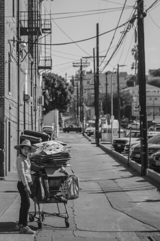 A woman stands with a shopping cart full of cardboard on a city street, captured in black and white.