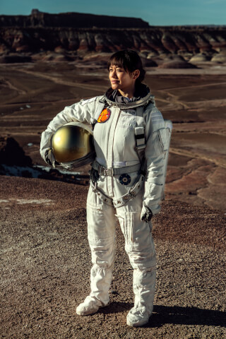 A female astronaut stands on a desert-like planet, holding a golden helmet.