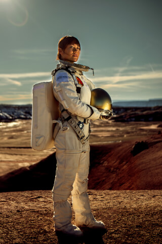 An astronaut stands on a mars-like planet, holding a helmet in her arms. The sun is rising in the background.