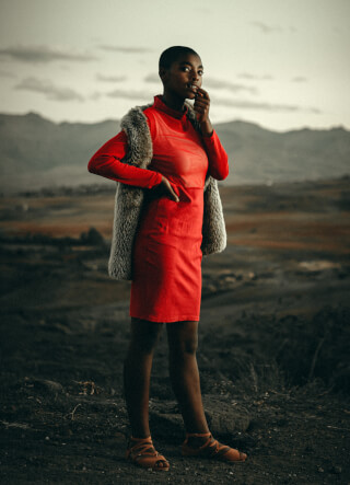A woman in a red dress and fur vest poses against a mountain backdrop.