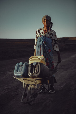 A woman hauls water cans in a wheelbarrow across a dry landscape, depicting water scarcity.