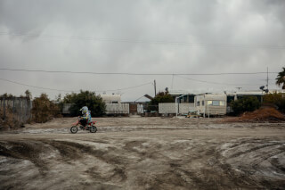 A child rides a dirt bike on a dusty road in a trailer park under a cloudy sky.