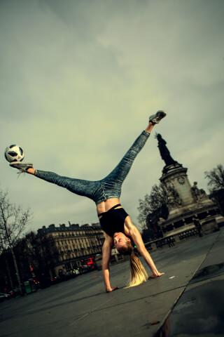 A woman performs a handstand with a soccer ball on her foot in an urban setting with a statue in the background.