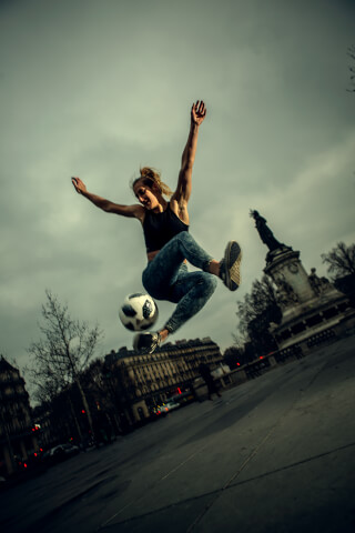 A female athlete jumps in the air, performing freestyle soccer tricks with a soccer ball in an urban setting.