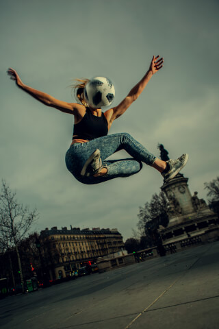 A woman performs soccer freestyle with a ball on her head against a backdrop of urban architecture.