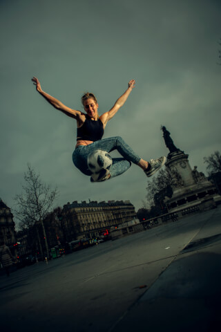 A woman performs freestyle football in an urban setting, demonstrating athleticism and skill in a dynamic action shot.