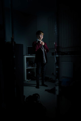 A young boy in school uniform adjusts his tie in his bedroom, illuminated by soft, ambient light.