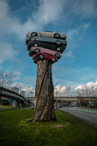 A whimsical sculpture featuring cars stacked atop a tree trunk, set against a backdrop of sky and cityscape.