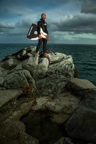 A man in a wetsuit stands on rocks by the sea, holding fins, under a cloudy sky, portraying a fitness and adventure lifestyle.