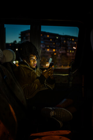A woman with dreadlocks uses her phone inside a vehicle at night with city lights in the background.