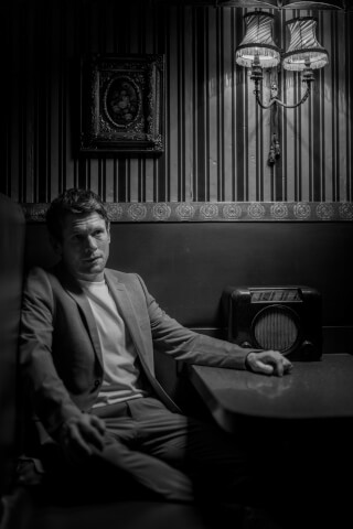 A man sits in a vintage booth, wearing a suit with a radio on the table in black and white.