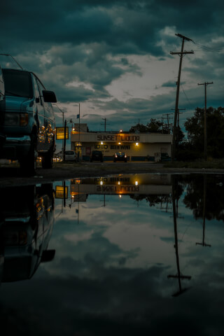 A nighttime shot of Sunset Liquor reflected in a puddle on a city street with a dark, moody atmosphere.