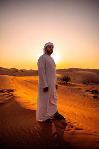 A man in traditional Arab dress stands in the desert at sunset.