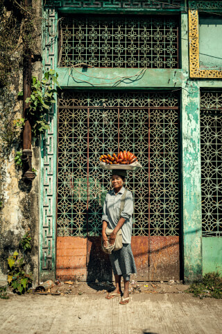 A woman carries fruit on her head in front of a weathered building, showcasing a scene of daily life.