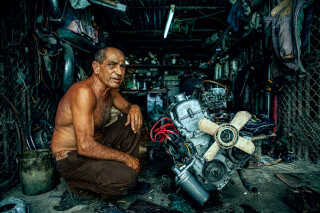 A mechanic crouches beside a disassembled engine in a cluttered workshop, focusing on his work.