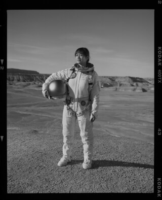 A woman in a spacesuit stands in a desert landscape, holding a helmet in this evocative black and white image.